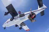 Two skydivers in tandem freefalling from a small airplane, with a third diver following closely. The plane's propellers are visible, and the sky is clear and blue. Both divers are wearing helmets and parachute gear.