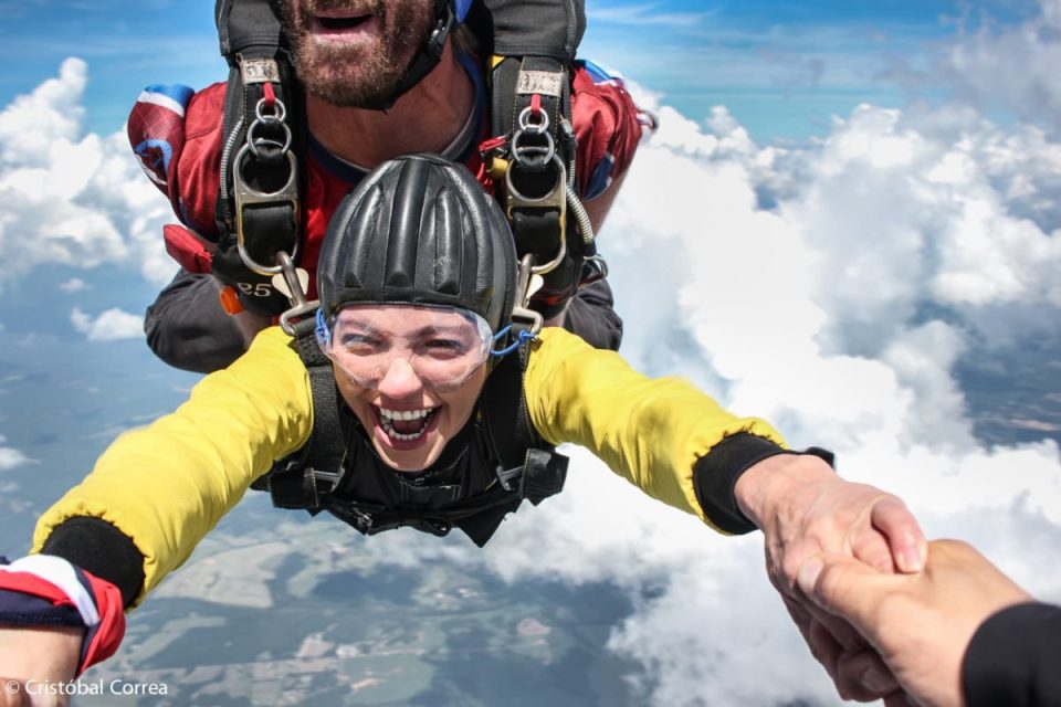 A person skydiving with an instructor, smiling broadly as they descend in tandem. The sky is clear with scattered clouds, and the person holds onto someone's hand creating a sense of excitement and adventure.