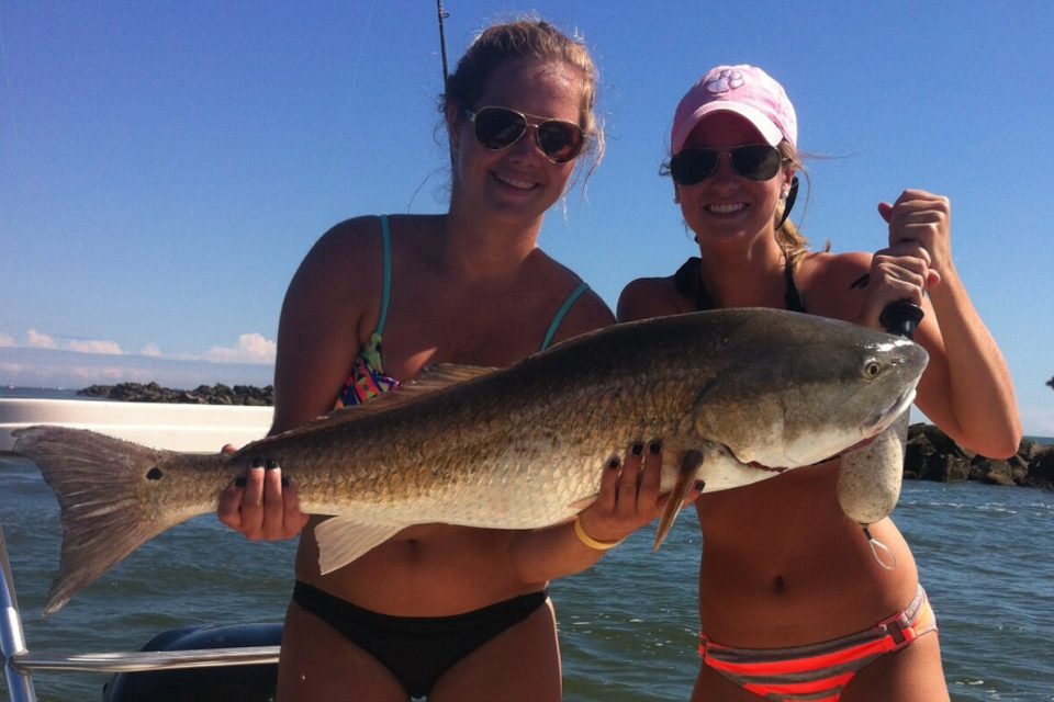 Two people on a boat holding a large fish, smiling at the camera. They are wearing swimwear and sunglasses. The sky is clear and the sea is calm.