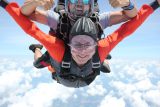 Two people tandem skydiving against a backdrop of blue sky and clouds. The person in front, wearing a helmet and goggles, appears excited. The instructor behind smiles broadly and gives a thumbs up. They are in freefall, experiencing the thrill of the jump.