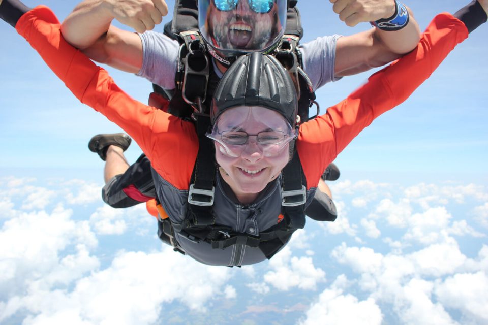 Two people tandem skydiving against a backdrop of blue sky and clouds. The person in front, wearing a helmet and goggles, appears excited. The instructor behind smiles broadly and gives a thumbs up. They are in freefall, experiencing the thrill of the jump.