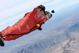 A person in a red wingsuit with goggles and a helmet skydives through a clear blue sky. The landscape below shows mountains, roads, and fields.