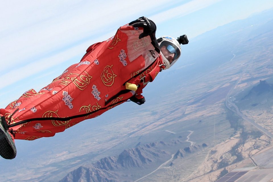 A person in a red wingsuit with goggles and a helmet skydives through a clear blue sky. The landscape below shows mountains, roads, and fields.