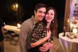 A smiling couple embraces at an outdoor evening event. The woman stands in front of the man, who hugs her from behind. They are surrounded by decorative lights and a table with bottles and a candle in the background.