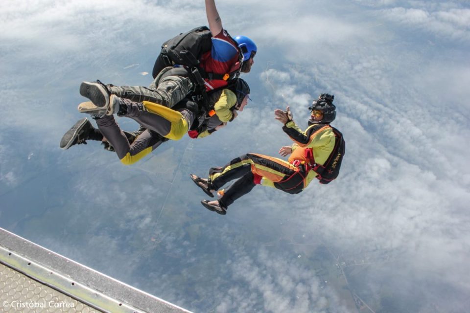 Three skydivers in mid-air, wearing colorful jumpsuits and helmets, against a backdrop of clouds and blue sky. One skydiver appears to be waving or gesturing at the others.