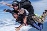 Two people are tandem skydiving through the clouds. The person in front is smiling and waving, wearing a black jumpsuit and goggles. The instructor behind is wearing a helmet and sunglasses. The sky is clear with scattered clouds below.