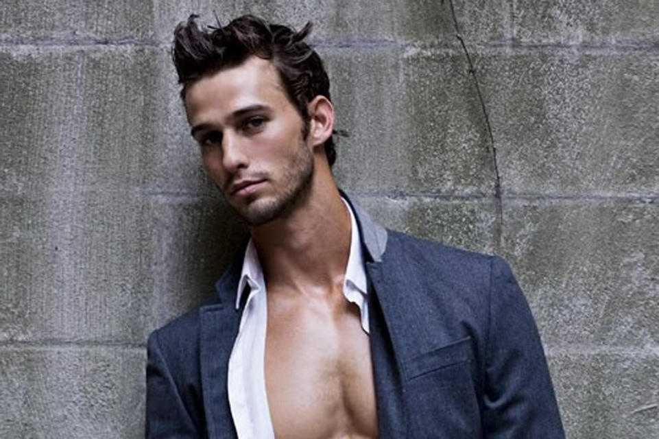 A man with short dark hair stands against a textured concrete wall. He is wearing a gray blazer over a partially unbuttoned white shirt, looking directly at the camera with a serious expression.