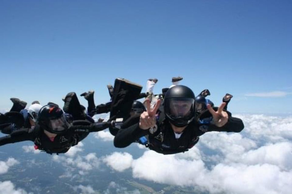 Two skydivers in freefall above the clouds, wearing black jumpsuits and helmets. One skydiver is making peace signs with both hands while smiling towards the camera. The sky is clear and blue with fluffy clouds below them.