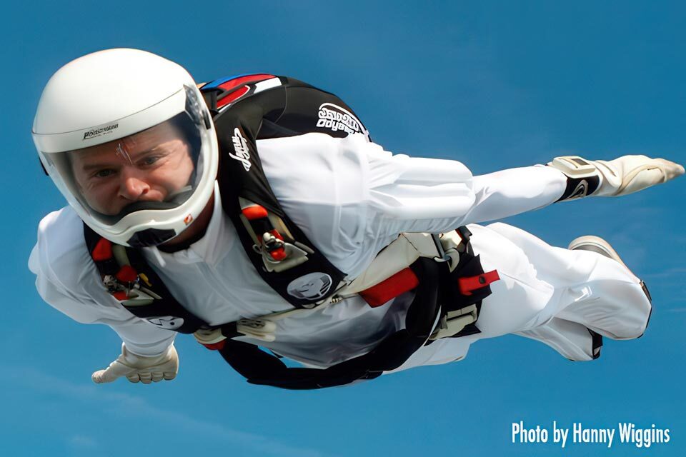 Skydiver in a white suit and helmet freefalling against a clear blue sky, with parachute gear visible on their back. Photo credit to Hanny Wiggins in the bottom right corner.