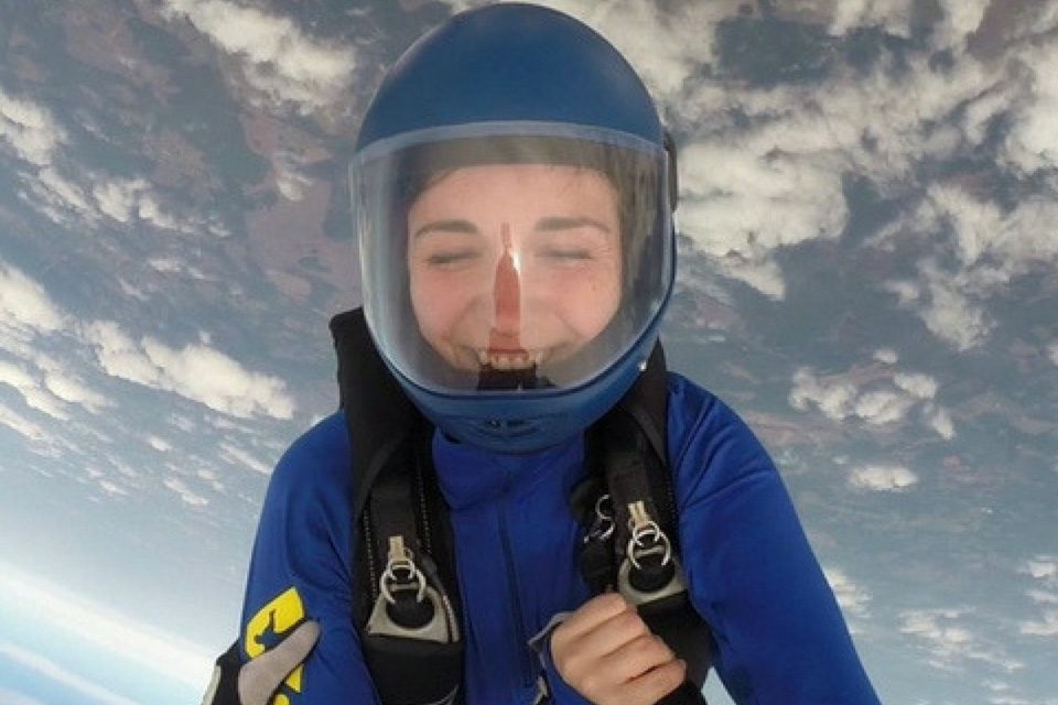 A person in a blue skydiving suit and helmet smiles broadly while skydiving. They are captured mid-air with a view of clouds and the earth beneath them.