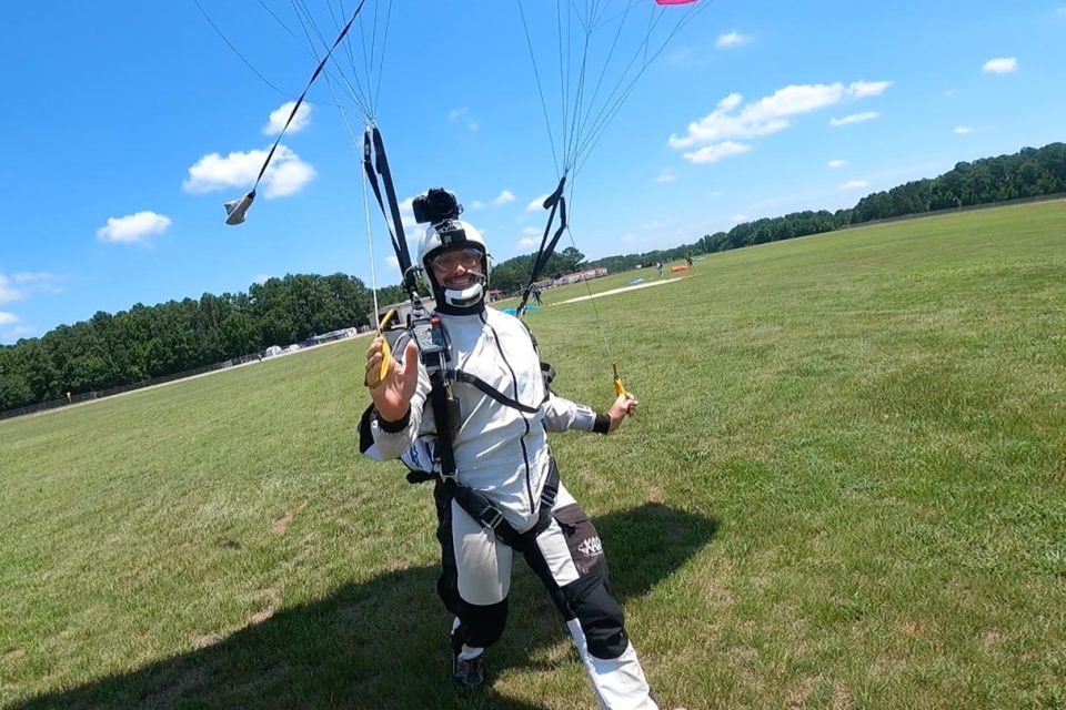 A person in a white and black skydiving suit lands on a grassy field with a parachute. They are holding a yellow object in each hand and smiling, wearing a helmet with a camera attached. A clear blue sky and trees are in the background.