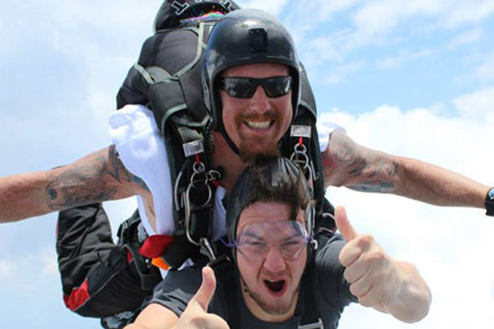 Two people in tandem skydiving gear free-fall through the sky. The person in front gives a thumbs-up, wearing goggles and a helmet. The person behind, also in a helmet, smiles while holding onto the harness. The sky is bright and partly cloudy.