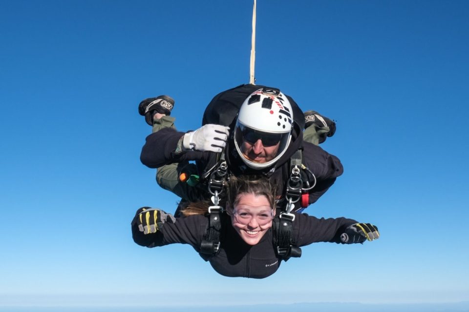 Two people are tandem skydiving against a clear blue sky. The front person smiles with arms outstretched, while the instructor behind them stabilizes the descent. Both wear protective gear, including helmets and goggles.