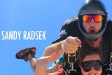 Two people skydiving, one wearing a helmet and sunglasses with their mouth open in excitement. The other person gives a thumbs up and wears sunglasses. Blue sky in the background with the name "Sandy Radsek" on the image.