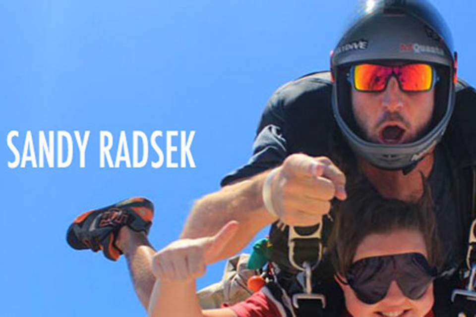 Two people skydiving, one wearing a helmet and sunglasses with their mouth open in excitement. The other person gives a thumbs up and wears sunglasses. Blue sky in the background with the name "Sandy Radsek" on the image.