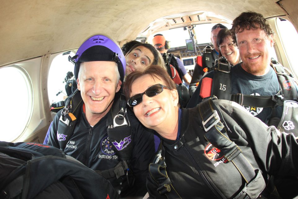 A group of six skydivers dressed in jumpsuits and wearing parachute gear are smiling inside a small aircraft, ready for a jump. They appear excited and cheerful as they pose for the camera.