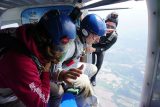 Three people in skydiving gear sit at the open door of an airplane. Two are equipped with helmets and parachutes, ready to jump. The sky and landscape are visible below. One person is in a crouched position, appearing ready to lead the jump.