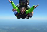 Two people are tandem skydiving against a clear blue sky. The person in front is smiling broadly, wearing a green and blue jumpsuit with outstretched arms. The instructor behind is guiding them with a focused expression.