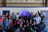 A large group of people, some in skydiving gear, pose together under a circular mural with signatures. They're inside a building with a sign above them. Many are smiling and pointing towards the center of the group.