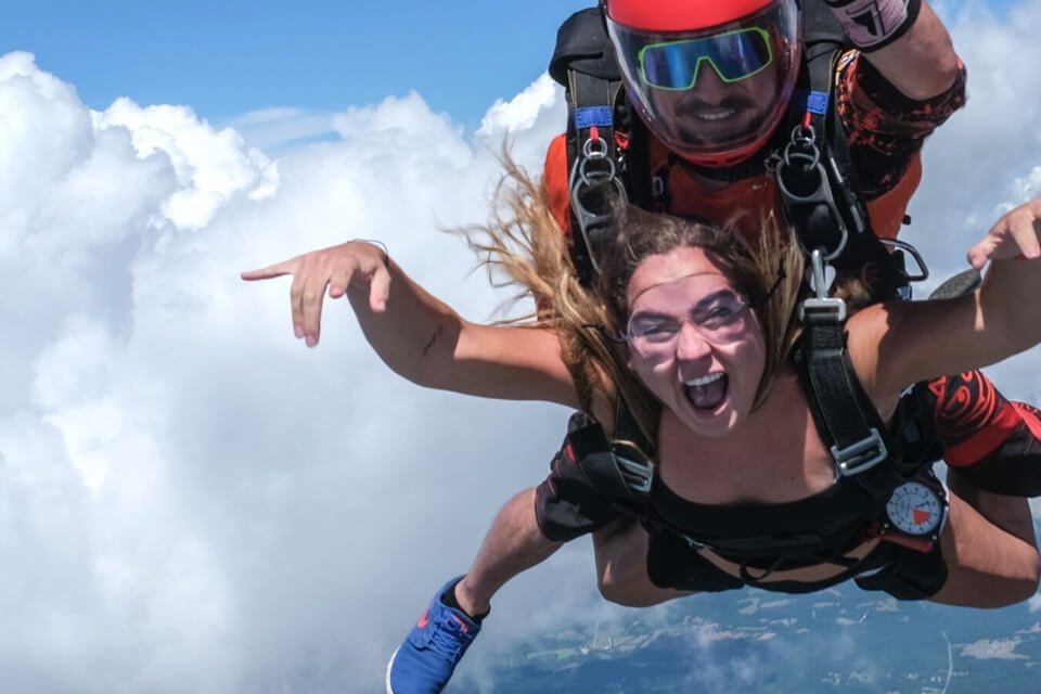 A woman and her instructor tandem skydiving through clear skies, with white clouds in the background. She has an excited expression, while the instructor wears sunglasses and a helmet. Both are harnessed together during the free fall.