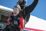 A woman skydiver, wearing goggles and a red and black jumpsuit, is smiling widely as she tandem skydives with an instructor. The sky is clear and blue, with part of the airplane visible in the background.