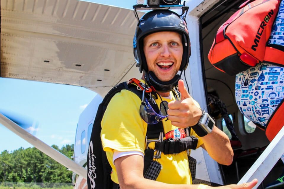 A person in a yellow shirt and helmet, giving a thumbs up, stands near an open aircraft door. Another person in a blue jumpsuit is boarding the plane. Both are wearing parachutes. The sky is clear in the background.