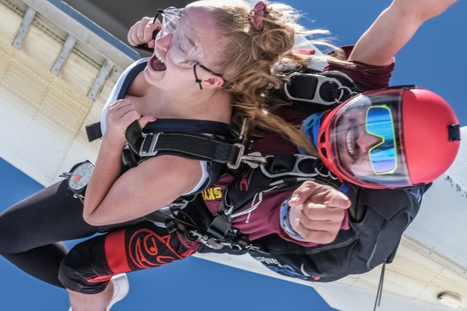 A tandem skydiving team exits a plane, with a woman strapped to an instructor. Both are wearing parachute harnesses and goggles. The instructor wears a red helmet and points forward, while the woman appears excited, against a clear blue sky.