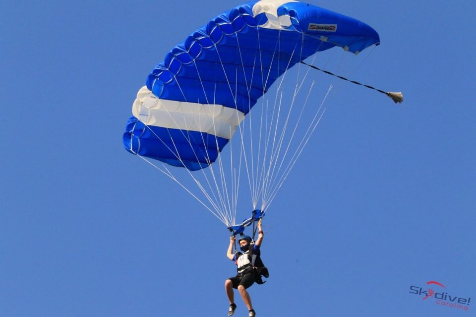 A person is skydiving under a blue and white parachute against a clear blue sky. The parachute has a "Skydive Carolina" logo on it.