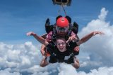 Two people tandem skydiving in the sky, surrounded by fluffy white clouds. The person in front is smiling broadly, wearing goggles, while the instructor behind wears a red helmet and sunglasses. They appear to be mid-fall, enjoying the thrilling experience.