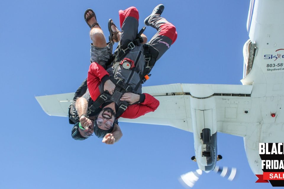 Two people in skydiving gear jumping out of an airplane, with one wearing goggles and a helmet. They're in a tandem skydive position against a clear blue sky. A "Black Friday Sale" banner is in the bottom right corner.