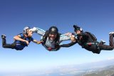 Three skydivers in freefall hold hands mid-air against a clear blue sky. They wear helmets and jumpsuits. The landscape below includes hills and a distant shoreline.