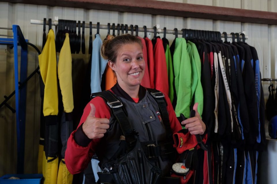 A person smiling and giving two thumbs up while wearing a skydiving harness. They are standing in front of a rack of colorful jumpsuits.