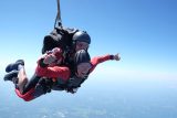 Two people skydiving in tandem, both wearing helmets and goggles. The person in front is smiling widely and giving a thumbs-up. The sky is clear and the landscape below is visible in the distance.