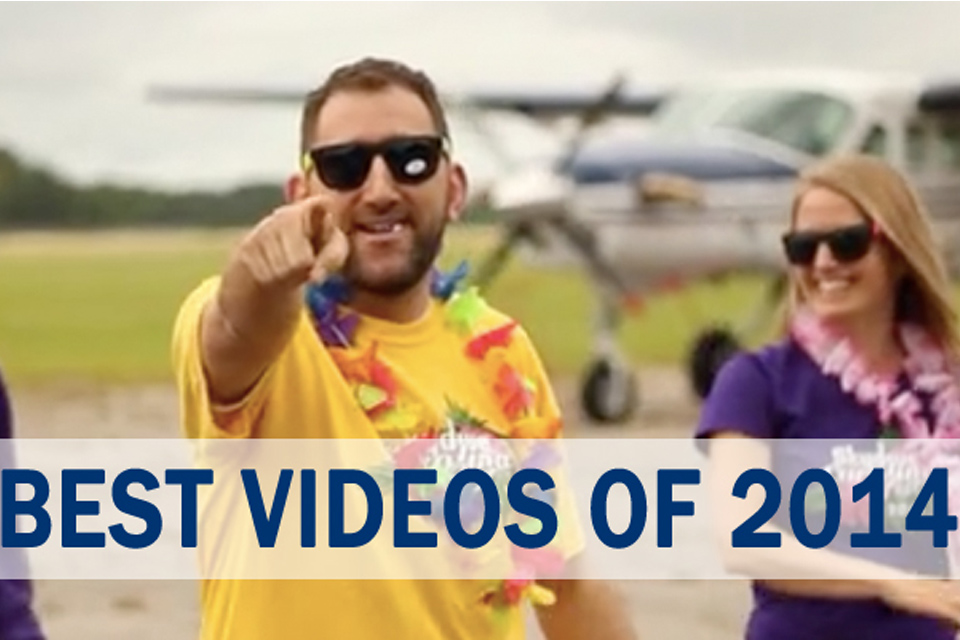 A man in sunglasses and a yellow shirt points at the camera while standing in front of a small airplane. A woman in a purple shirt and sunglasses stands beside him, both wearing colorful leis. Text overlay reads "BEST VIDEOS OF 2014.