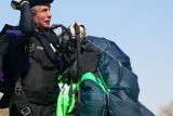 A man in a black jumpsuit and helmet is holding a parachute. The parachute is partially packed, with bright green and blue panels visible. He is outdoors under a clear blue sky.