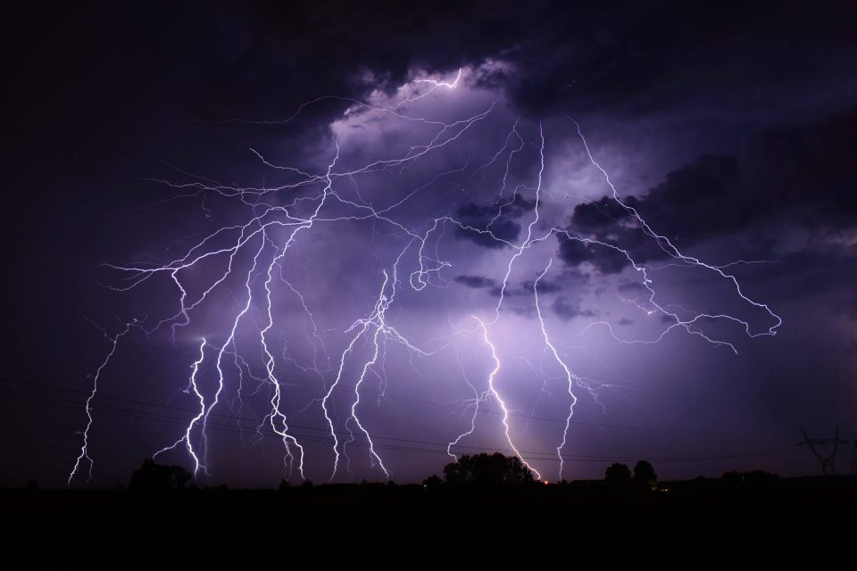 A dramatic night sky filled with multiple streaks of lightning illuminating the dark clouds. Silhouettes of trees and power lines are visible at the bottom against the bright, electric light.