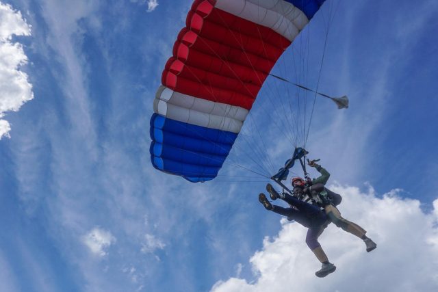 Two people tandem skydiving under a red, white, and blue parachute against a partly cloudy sky. The parachutists are descending with legs stretched forward, enjoying the flight.