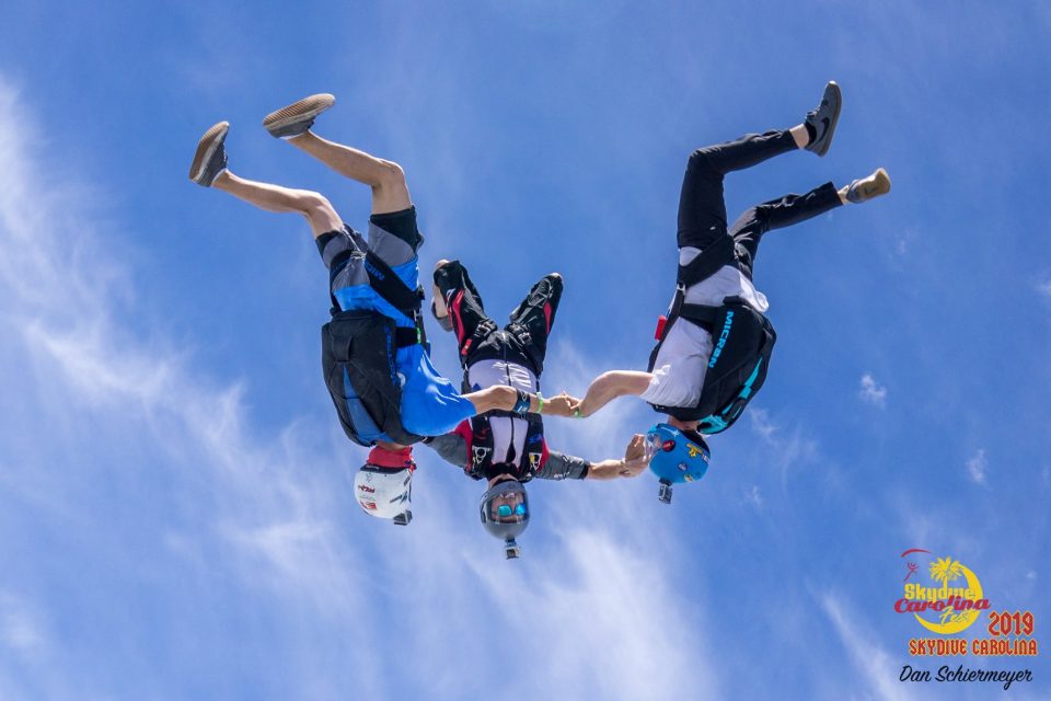 Three skydivers in freefall hold hands mid-air against a backdrop of a clear blue sky. Each wears a helmet and parachute. The logo "Skydive Carolina 2019" is visible in the bottom right corner.