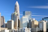 A city skyline with a cluster of modern skyscrapers, including a prominent building with a pointed top. The sky is clear and blue, enhancing the buildings' glass reflections.