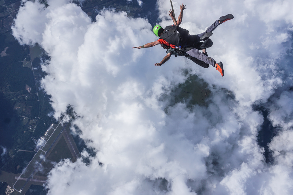 Two skydivers in tandem freefall through the sky, surrounded by thick, white clouds. One diver wears a bright green helmet. Below, an airstrip and forested landscape are visible through gaps in the clouds.