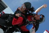 Two individuals are tandem skydiving, wearing helmets and jumpsuits, with one person's face visible and displaying an expression of excitement or thrill. The sky is clear and blue in the background.