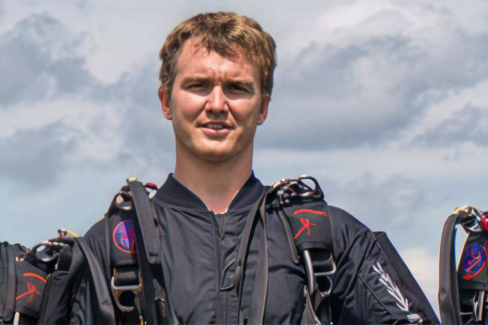 A person in a black jumpsuit stands outdoors against a cloudy sky, wearing a parachute harness. The harness features various patches and straps. The scene suggests preparation for a skydiving activity.