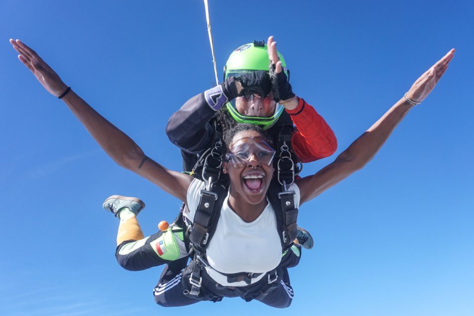 Two people tandem skydiving against a clear blue sky, wearing harnesses and gear. The person in front is smiling with arms outstretched, while the instructor behind them manages the jump. Both are wearing goggles.