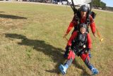 Two people in red jumpsuits land on a grassy field after a tandem skydive. The person in front is seated with legs extended, while the instructor behind supports them. Both are wearing helmets and harnesses, smiling and appearing excited.