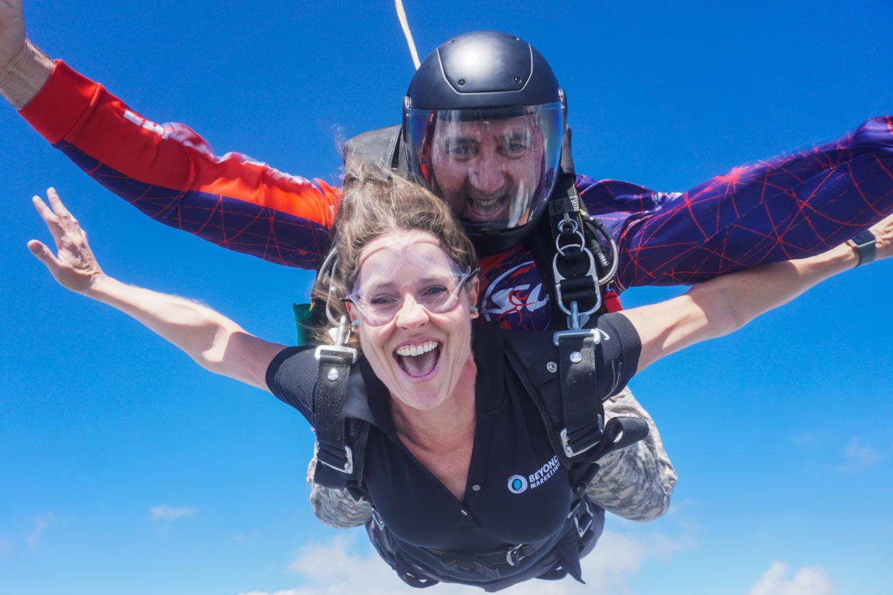 A woman and her tandem skydiving instructor are free-falling through the sky. The woman, wearing glasses, smiles widely, with her arms outstretched. The instructor is behind her, wearing a helmet and sunglasses, under a clear blue sky.