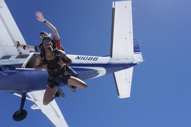 A tandem skydiving pair exits an airplane against a clear blue sky. The instructor is wearing a jumpsuit, helmet, and goggles, while securely holding the excited diver. The plane, painted white and blue, is visible in the background.