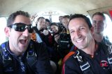 A group of smiling people in skydiving gear sit closely inside an airplane. Some are wearing sunglasses, and one holds a camera. They appear excited, possibly preparing for a skydive. The interior of the plane is visible around them.