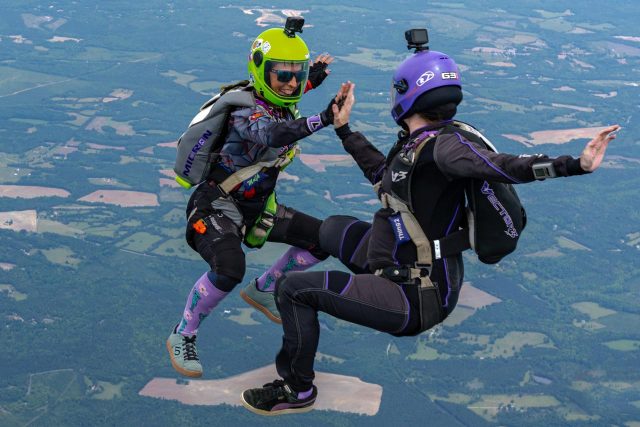 Two skydivers wearing helmets and specialized suits perform a mid-air high-five. They are free-falling above a landscape of fields and forests. Both are equipped with action cameras on their helmets, capturing the exhilarating moment.