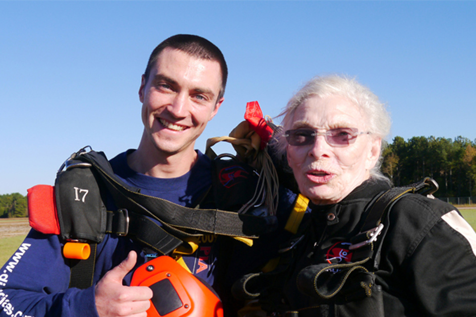 A smiling man and an older woman wearing skydiving gear stand together outdoors under a clear blue sky. They appear happy and excited.