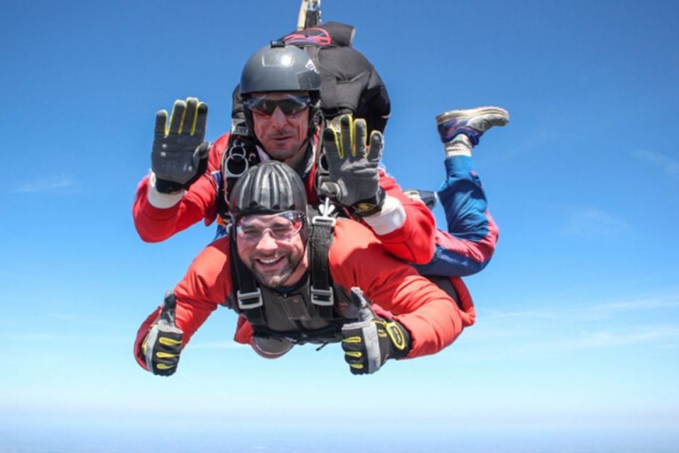 Two people tandem skydiving against a clear blue sky. The person on top is guiding with a parachute pack, both wearing helmets and goggles, smiling as they descend. The front person wears a red suit and gloved hands are visible in the foreground.